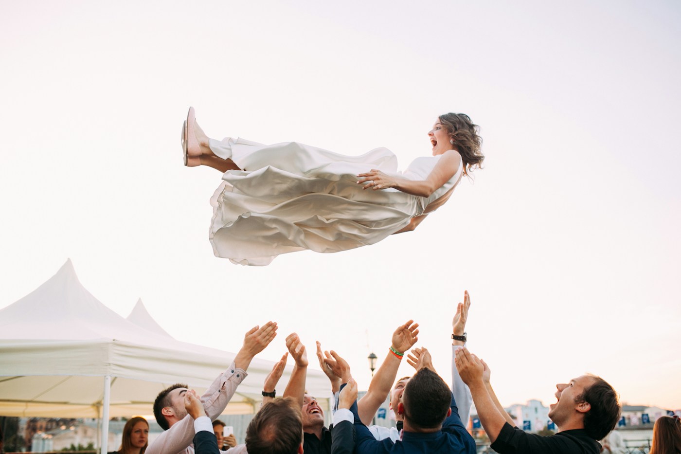 Guests tossing a laughing bride into the air at an outdoor wedding